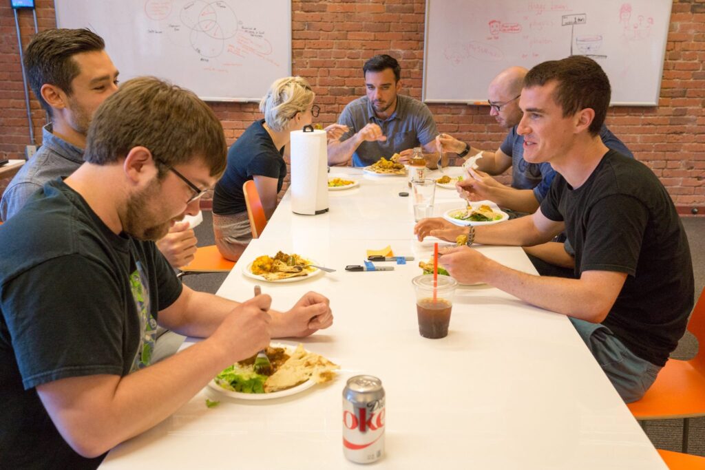 Co-workers talking and enjoying office group lunch from Lish at a long table with a plate of food and drinks.
